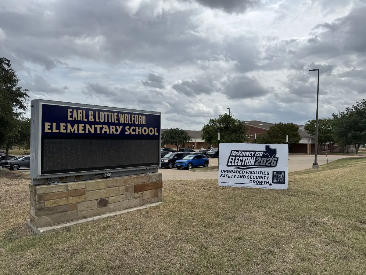 A white McKinney ISD Election 2026 sign on the lawn beside the Earl and Lottie Wolford Elementary School sign, with the school building and a parking lot behind.